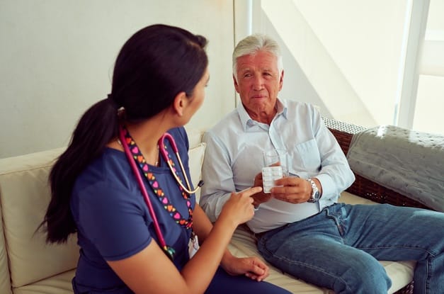 A nurse sitting on the couch with an older gentlemen. She is pointing to a white pack of pills in his hand.