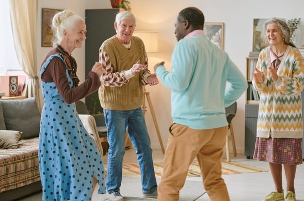 Four diverse senior individuals dance and socialize in the center of a living room at a senior nursing home.