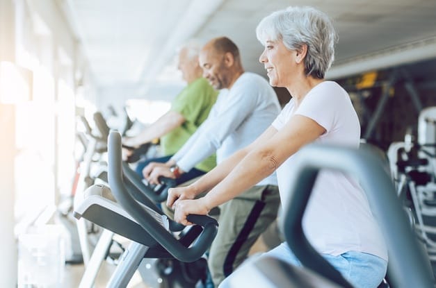 A group of older people sitting on exercise bikes. There is a woman with gray hair and a white shirt using one of the bikes.