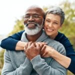 A smiling, gray-haired woman hugs a man from behind. The man holds her hands and has his eyes closed.