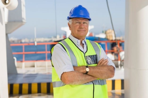 A man with white hair and eyeglasses, wearing a blue hard hat and a bright yellow safety vest, crosses his arms.