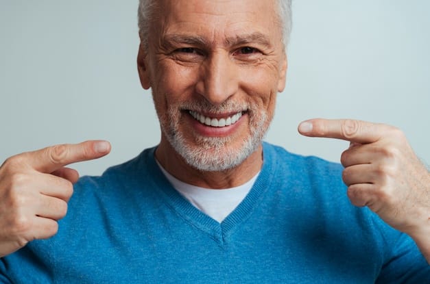 A senior adult wearing a blue sweater has a large smile on his face. He is pointing to his smile with both hands.