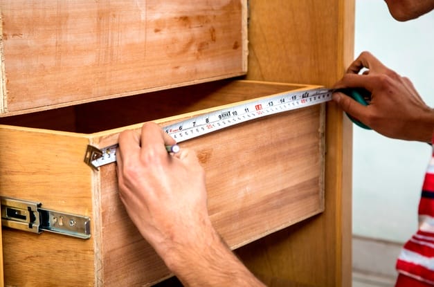 A close-up of a man using a tape measure to mark the length of the front of a half-pulled-out wooden drawer on a dresser.