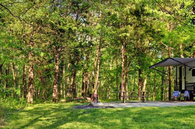 A camper sits on a concrete slab that is surrounded by large, mature trees. Two folding chairs sit next to the camper.