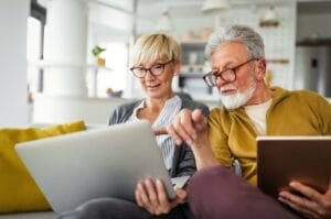 A senior man and woman wearing glasses sitting on a couch in a brightly lit living room using a tablet and a laptop.