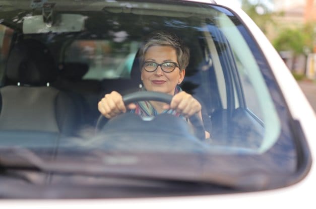 An older woman wearing glasses sits behind the wheel of a car, driving somewhere. She has both hands on the wheel.