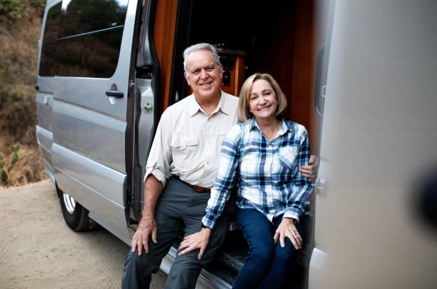 A smiling older couple dressed in button-up shirts sit in the doorway of their silver camper van.