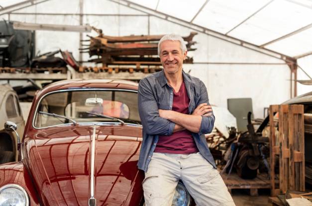 An older man sitting on the hood of a polished classic car and smiling. There are pallets and other equipment around the car.
