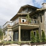 Cloudy skies stretch over a green three-story house with glass balconies, wood furniture, and a stone chimney.