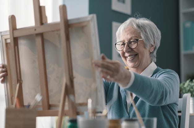 A smiling senior woman holds her arms out to admire her painting that rests on a wooden easel.