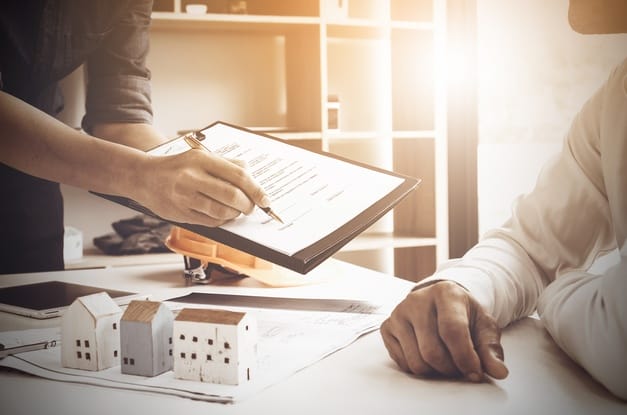 One person holds a pen and a document out toward another person. Models of properties sit on papers on top of a desk.