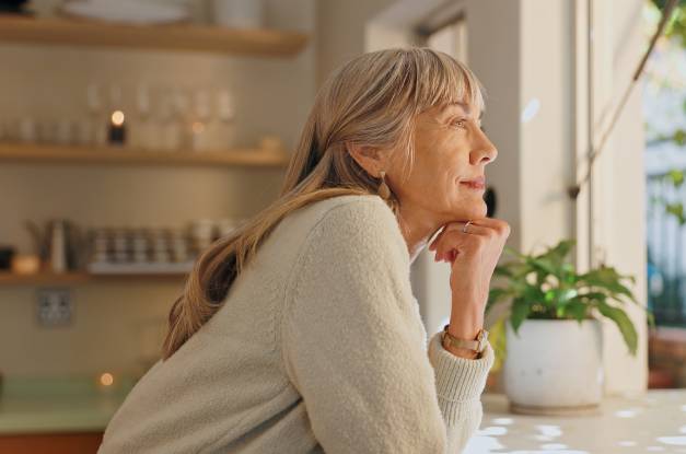 A senior woman is leaning forward on a counter with her face resting on her knuckles, looking out a window.
