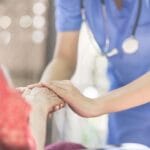A nurse in blue scrubs bends over slightly to gently support and cover the hands of a seated elderly woman.