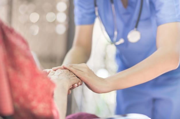 A nurse in blue scrubs bends over slightly to gently support and cover the hands of a seated elderly woman.