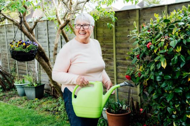 A smiling senior woman stands holding a green watering can in her backyard next to a fence and various plants.