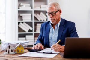 An older man sits at a desk in front of a laptop, writing on a piece of paper and using a calculator.