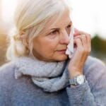 A white-haired woman in a gray top and scarf wipes her face with a tissue while in front of a blurred background.