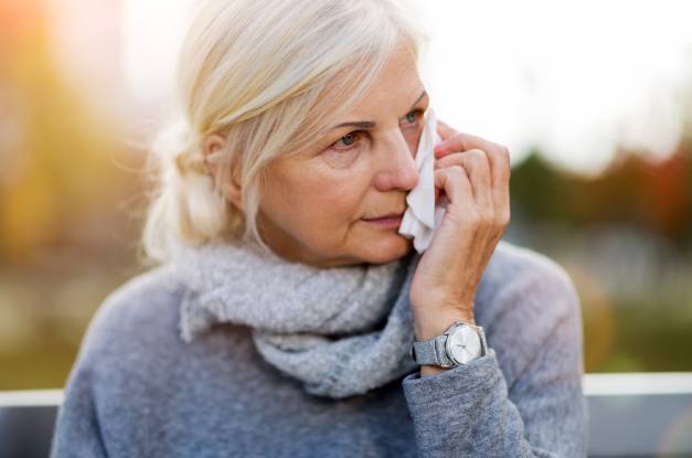 A white-haired woman in a gray top and scarf wipes her face with a tissue while in front of a blurred background.
