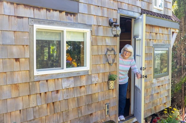 A woman in a striped shirt and jeans leans out the front door of a small wooden house with a cheerful expression.