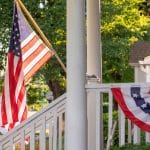 A large American flag hangs off the end of a white flagpole attached to a pillar on the front porch of a home.