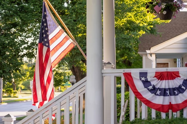 A large American flag hangs off the end of a white flagpole attached to a pillar on the front porch of a home.