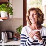 A smiling senior woman in a striped shirt holding a coffee cup inside a sunny kitchen filled with plants.