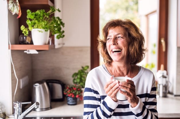 A smiling senior woman in a striped shirt holding a coffee cup inside a sunny kitchen filled with plants.