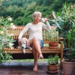 An older woman sits cross-legged on a bench while sipping coffee next to a dog, surrounded by potted plants.