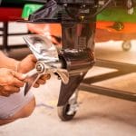 A man kneels down in front of his boat, holding a wrench to the boat's propeller to replace it before winter.