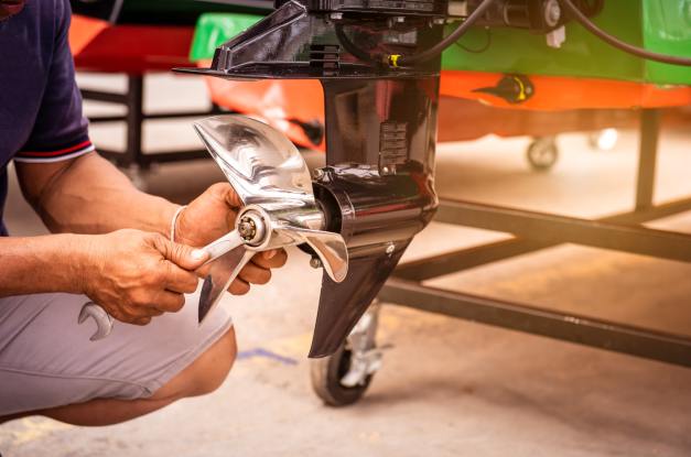 A man kneels down in front of his boat, holding a wrench to the boat's propeller to replace it before winter.