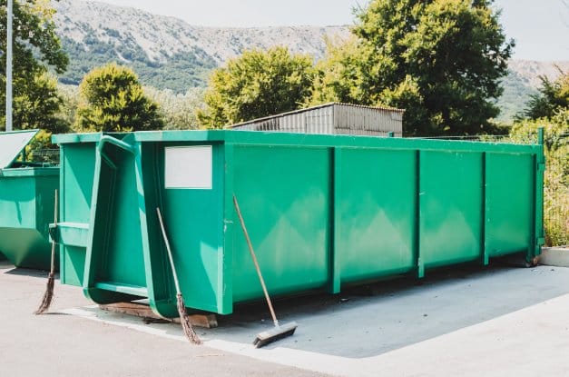 A bright green large waste container sits on concrete. Brooms lean on the container and behind it are the mountains.