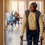 An elderly man walking out of a room with the help of a walker. He has both hands on the handles of the mobility aid.