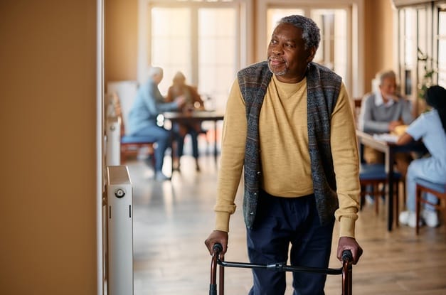 An elderly man walking out of a room with the help of a walker. He has both hands on the handles of the mobility aid.