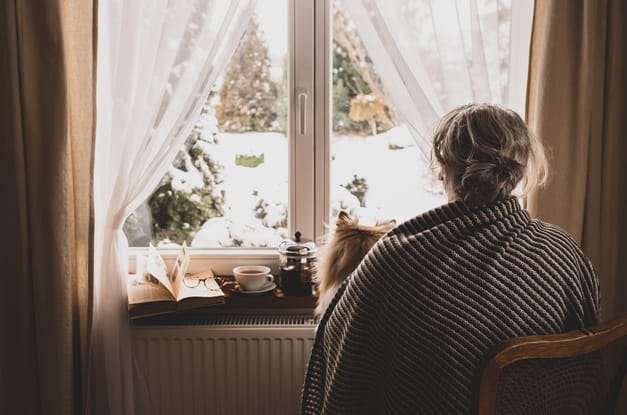 A woman sits with her dog, looking out the window at her yard, which is covered in snow. She wears a blanket to keep warm.