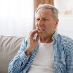A man sitting on a couch, holding his cheek and clenching his teeth, with a pained expression on his face.