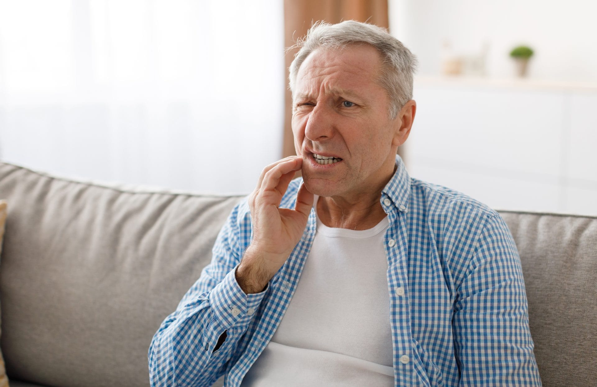 A man sitting on a couch, holding his cheek and clenching his teeth, with a pained expression on his face.