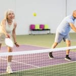 An older woman and a man are playing pickleball. The older woman is about to hit the ball with her right hand.