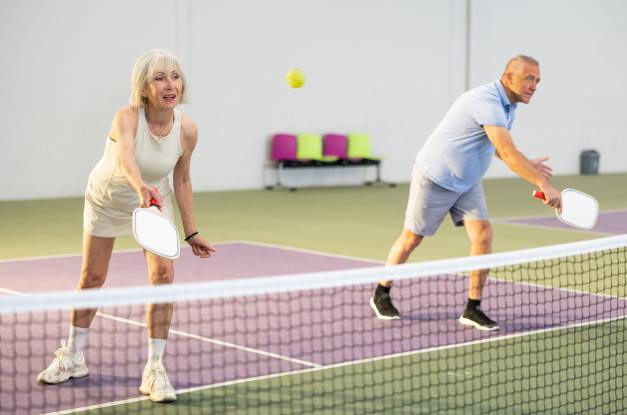 An older woman and a man are playing pickleball. The older woman is about to hit the ball with her right hand.