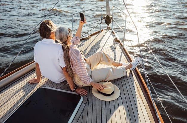 A senior couple sits on a sailboat deck at sunset as the woman takes a selfie of them during a romantic date on the water.