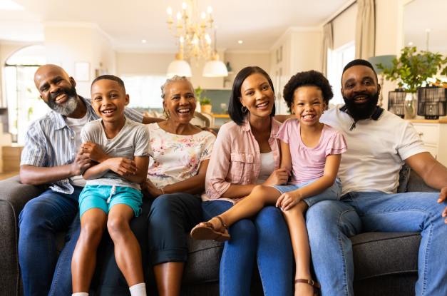 Three generations of a family sitting together and smiling on a couch in a home with a kitchen in the background.