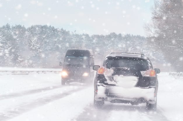 Two cars are driving on opposite sides of the road during a snowstorm. The rear of one car is covered in snow.