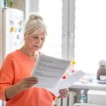 Home 10 A senior adult woman stands in a bright kitchen, holds two sheets of paper, and reads the information.