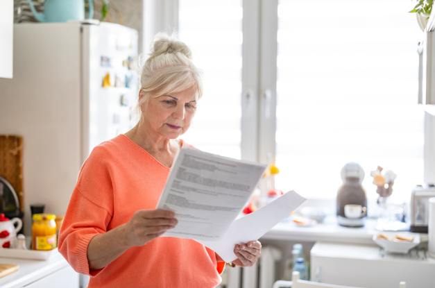 A senior adult woman stands in a bright kitchen, holds two sheets of paper, and reads the information.