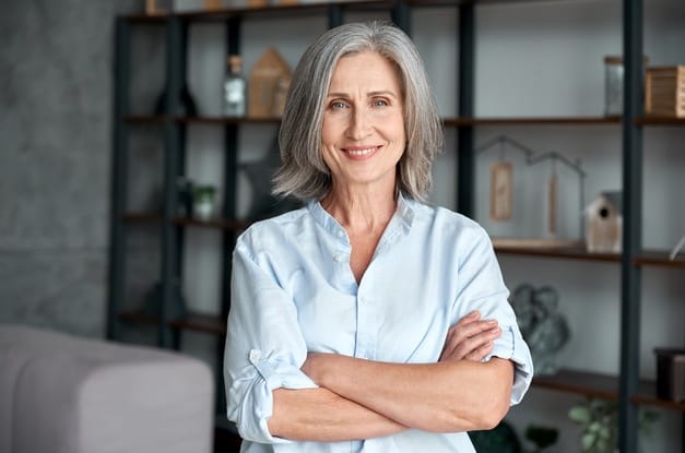 A woman stands with her arms folded as she looks at the viewer and smiles. A decorative bookshelf is behind her.