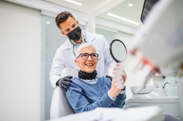 An older woman with white hair sits in a dental exam chair and smiles into a handheld mirror with a dentist standing behind her.