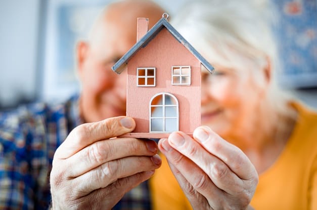 A small painted wooden model of a home getting held up by a blurred senior couple smiling in the background.