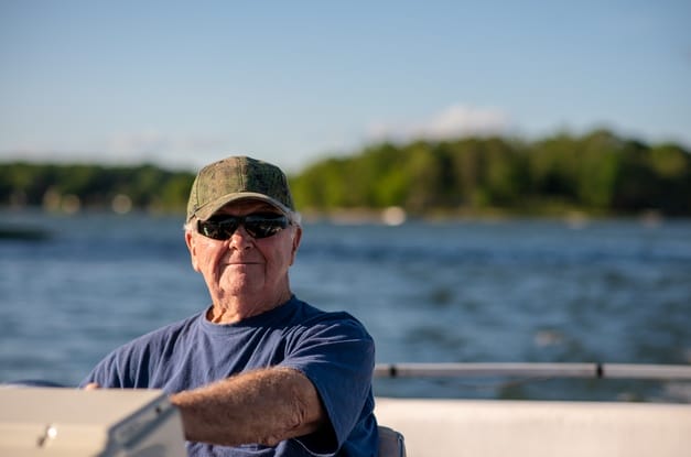 A senior man in a blue T-shirt, green baseball cap, and sunglasses sits at the helm of a boat on a lake.