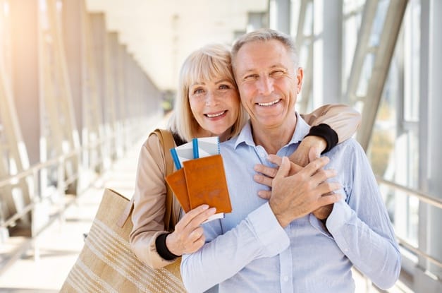 A couple embraces while holding their plane tickets and standing in an airport. Bright sunlight fills the setting.
