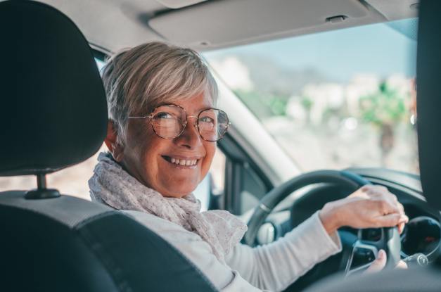 An older woman smiles as she sits behind the wheel of her car. She wears glasses and has her hand on the steering wheel.